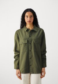 Olive green, button-up shirt made of sturdy fabric, featuring two chest pockets and a fold-over collar. Long sleeves, relaxed fit.