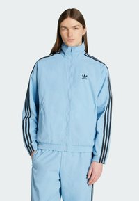 Lichtblauwe Adidas trainingsjack met zwarte strepen langs de mouwen, hoge kraag, ritssluiting aan de voorkant, zijzakken en elastische manchetten.