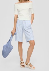 Haut blanc à épaules dénudées, short rayé bleu clair et blanc, sandales beiges épaisses, tenant un sac fourre-tout tissé bleu.