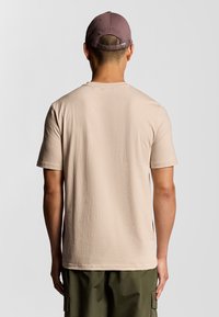 T-shirt di cotone beige con maniche corte, collo a giro, e vestibilità rilassata. Abbinata a un cappellino viola con marchio ricamato.