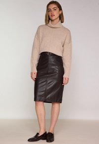 Pull en maille beige à col montant associé à une jupe crayon en cuir noir avec des coutures et une taille ceinturée. Des mocassins sombres complètent le look.