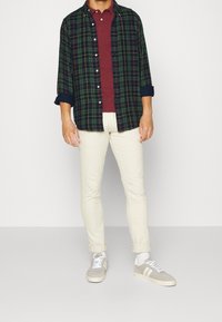 Plaid flannel shirt in dark green, navy, and red, layered over a maroon polo. Cream-colored slim-fit pants and gray sneakers with white accents.