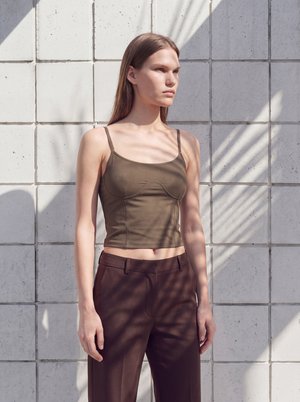 Γυναίκα με καφέ αμάνικο crop top και σκούρο καφέ παντελόνι στέκεται δίπλα σε πλακιδωτό τοίχο με σκιές από φύλλα φοίνικα.