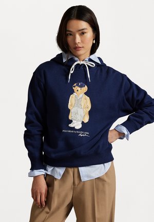 Felpa blu navy realizzata in cotone con un design di orsetto polo ricamato, dotata di un laccio bianco e polsini a costine, indossata sopra una camicia azzurra chiara.