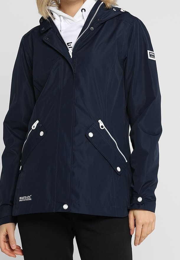 Femme portant une veste imperméable bleu marine avec fermeture éclair et poches pressionnées, un sweat à capuche blanc en-dessous, et un pantalon noir sur un fond uni.