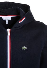 Sudadera con cremallera de color azul marino que presenta un logo de cocodrilo verde, acentos a rayas rojas y blancas en la capucha y una tela suave y texturizada.