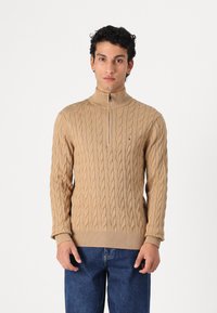 Beige cable-knit sweater with a high collar and quarter zipper. Features ribbed cuffs and hem, made from a soft textured material.