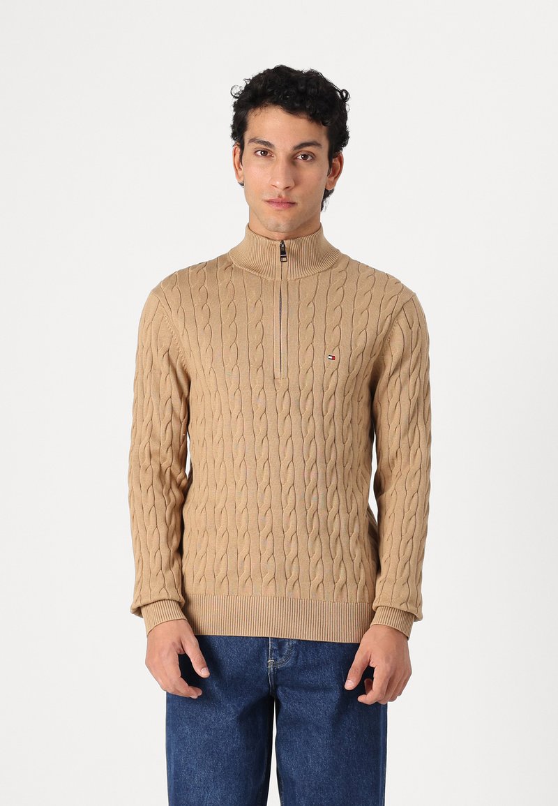 Beige cable-knit sweater with a high collar and quarter zipper. Features ribbed cuffs and hem, made from a soft textured material.