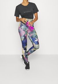 Maglietta nera cropped con logo, leggings floreali multicolori e scarpe da ginnastica nere. Tiene una bottiglia d'acqua trasparente rosa.