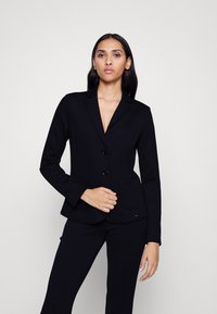 Blazer noir ajusté avec deux boutons, revers cranté et poches à rabat, fabriqué à partir d'un tissu lisse, associé à un pantalon noir assorti.
