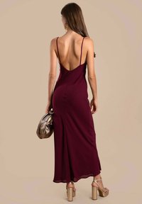 Burgunder midi Kleid mit einem tiefen Rücken, dünnen Trägern und einer fließenden Silhouette. Die Accessoires umfassen eine metallische Tasche und klobige Plattformabsatzschuhe.