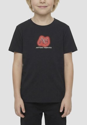 Niño con una camiseta negra con un logo "AM" rojo brillante y texto blanco "ANTONY MORATO" en el pecho, de pie frente a un fondo liso.