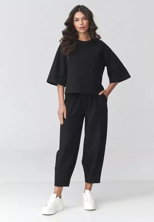 Kobieta z długimi kręconymi włosami, ubrana w luźną czarną bluzkę, czarne krótkie spodnie i białe masywne sneakersy, stoi na tle jednolitego tła.