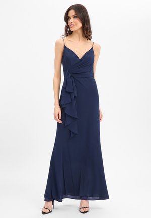 Frau steht und trägt ein dunkelblaues ärmelloses Abendkleid mit dünnen Trägern und Rüschendetail an der Seite, kombiniert mit schwarzen offenen Spitzenabsätzen.