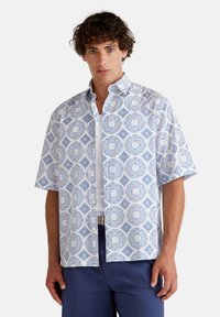 Camisa de manga corta de tela blanca con un patrón geométrico azul de círculos y cuadrados. Cuello abotonado y corte relajado.