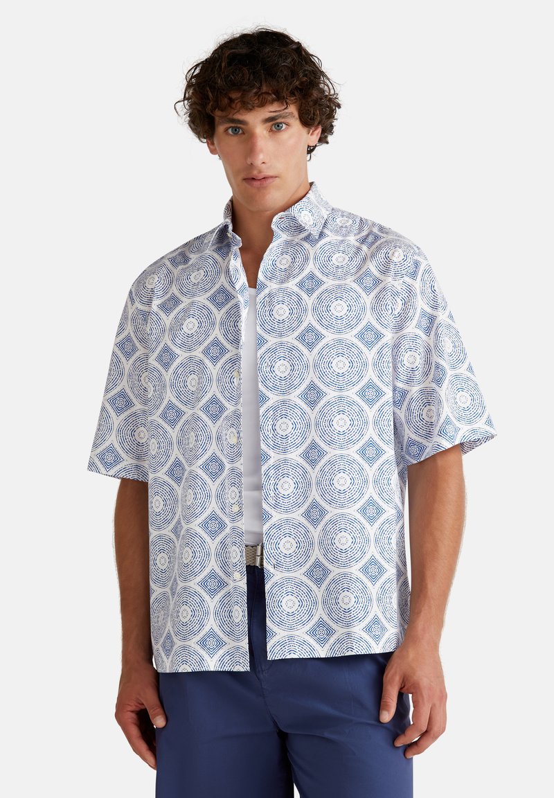 Camisa de manga corta de tela blanca con un patrón geométrico azul de círculos y cuadrados. Cuello abotonado y corte relajado.