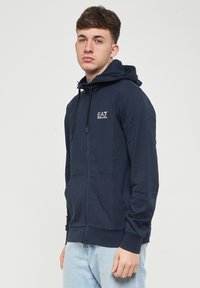 Felpa con zip color blu navy, dotata di tasche frontali, cappuccio regolabile con lacci e logo EA7 sul petto sinistro, realizzata in tessuto morbido e traspirante.