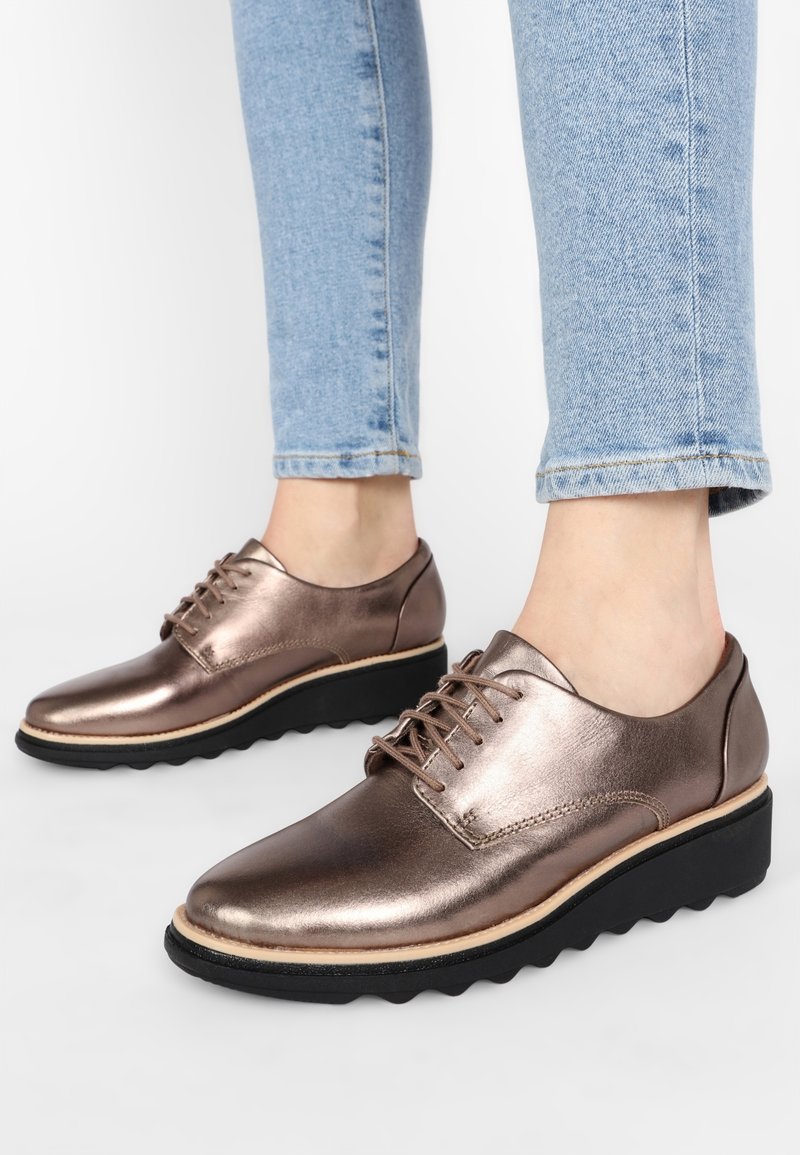 Bronzen metallic leren schoenen met een ronde neus, veterontwerp en een zwarte gesegmenteerde zool. Draagt lichte blauwe jeans van enkellengte.