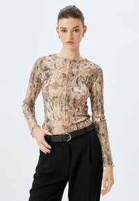 Langermige top met een slangenprint in beige en bruin, met een ronde halslijn en subtiele plooien bij de mouwen. Gecombineerd met zwarte broek en een riem.