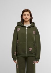 Olivinės spalvos zip-up bliuzonas iš minkštos medžiagos su didele gobtuvu, su rožinėmis siuvinėtomis logo ir priekinėmis kišenėmis. Derinamas su atitinkančiomis kelnėmis.