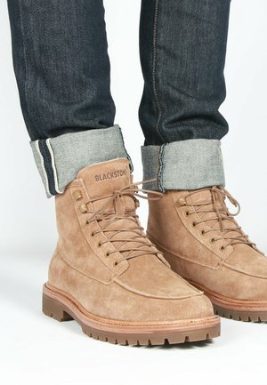 Botas de ante marrón con cordones y suela resistente, acompañadas de jeans oscuros remangados con un borde de mezclilla clara.