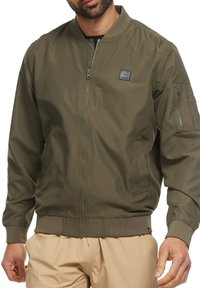 Homme portant une veste bomber vert olive avec zip sur le devant, poche zippée sur la manche, et pantalon beige, visible du cou aux hanches.
