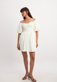 Off-shoulder vit klänning med puffärmar, rynkad bodice, rynkad kjol och slät textur. I kombination med svarta remsandaler.