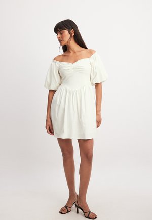 Off-shoulder witte jurk met pofmouwen, geraffineerde taille, geplooide rok en een gladde textuur. Gecombineerd met zwarte strappy hakken.