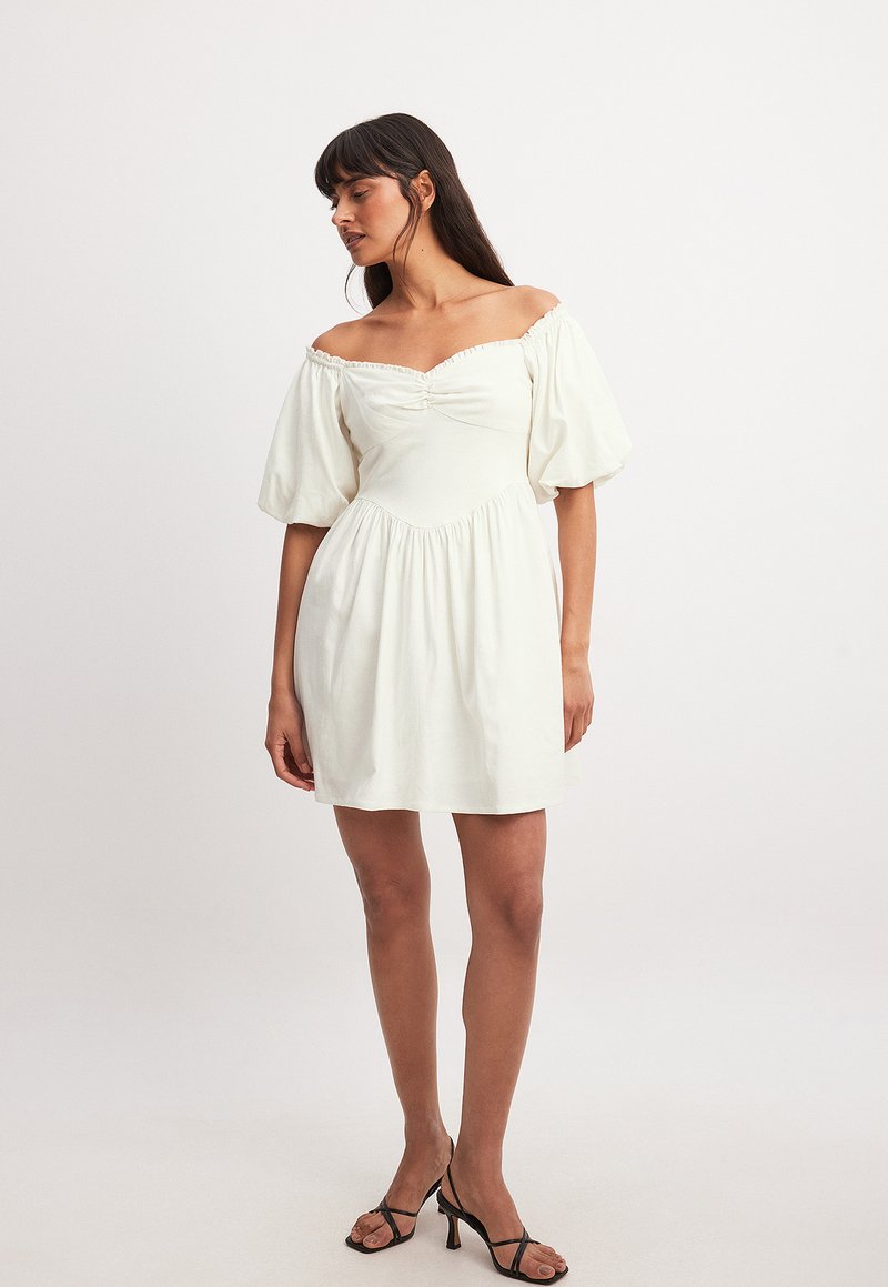 Off-shoulder vit klänning med puffärmar, rynkad bodice, rynkad kjol och slät textur. I kombination med svarta remsandaler.