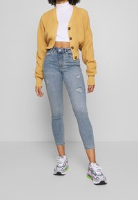 Cárdigan de punto amarillo con mangas oversized, cierre de botones, combinado con jeans ajustados desgastados de color azul claro y zapatillas grises con acentos verdes.