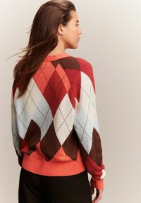 Femme aux longs cheveux bruns portant un pull en motif argyle rouge, marron, blanc et bleu clair, se tenant devant un fond beige uni.