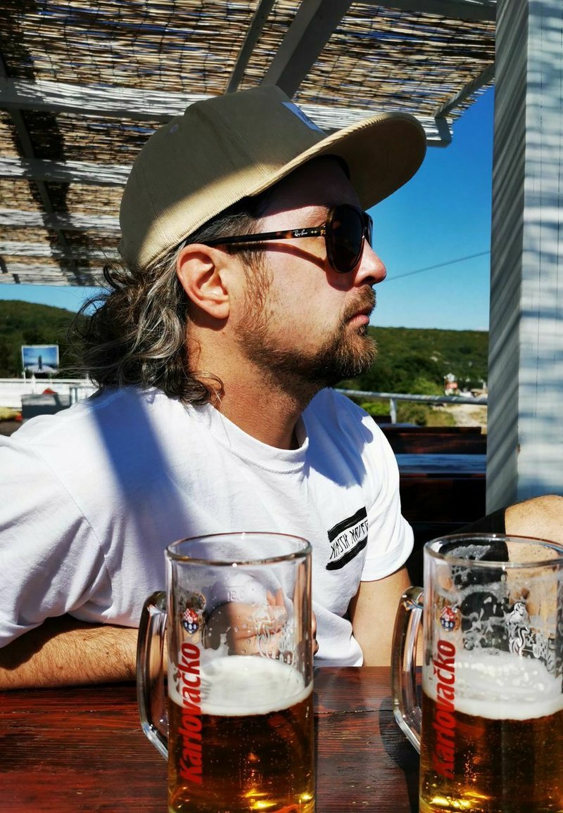 Man with long hair wearing a tan baseball cap and sunglasses, seated at a wooden table, with two large mugs of beer in front.