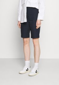 Persona vestida con pantalones cortos negros hasta la rodilla, camisa blanca de manga larga, calcetas blancas y zapatillas blancas con logos negros, de pie frente a un fondo liso.