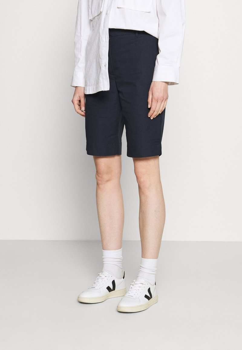 Persona vestida con pantalones cortos negros hasta la rodilla, camisa blanca de manga larga, calcetas blancas y zapatillas blancas con logos negros, de pie frente a un fondo liso.