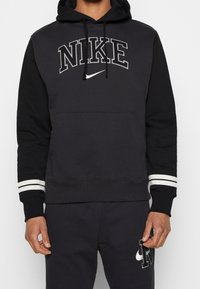 Svart Nike hoodie med grå text, känguruficka och randiga detaljer på ärmsluten. Materialet verkar mjukt och har en avslappnad passform.