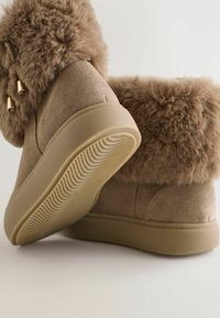 Botoși din suede maro cu bordură din blană sintetică, având o talpă din cauciuc texturat și accente metalice. Design robust cu o textură moale și pufoasă.