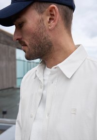 Bílá košile na knoflíky s límečkem, nošená přes bílý tričko. Výrazný detail loga na košili. Měkká tkanina s hladkou texturou.