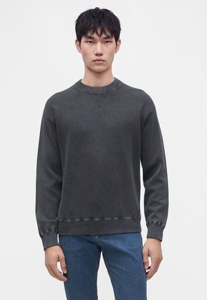 Ung mand iført en mørkegrå crewneck sweatshirt og blå jeans, stående mod en ensfarvet lys baggrund, vendt mod fronten.