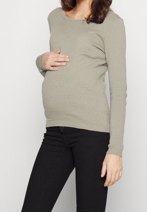 Femme enceinte portant un pull texturé gris clair et un pantalon noir, tenant doucement son ventre d’une main.