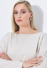 Junge Frau mit glattem blonden Haar und blauen Augen, die einen weißen Pullover mit Strasssteinen und silbernen Creolen trägt, mit verschränkten Armen.