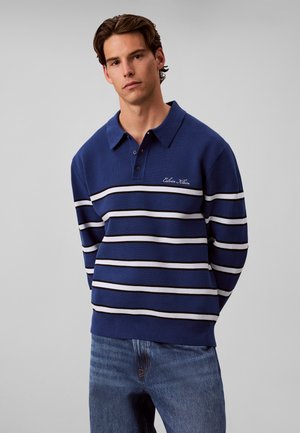 Jeune homme portant un polo bleu à manches longues avec des rayures horizontales blanches et noires et un jean bleu, debout devant un fond uni.