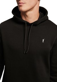Homme portant un sweat à capuche noir avec cordons de serrage et un petit logo blanc représentant une personne balançant un club de golf sur la poitrine.