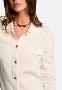 Cardigan en tricot crémeux avec des boutons dorés et des poches sur la poitrine, doté d'un col. Collier épais en or et noir en accent.