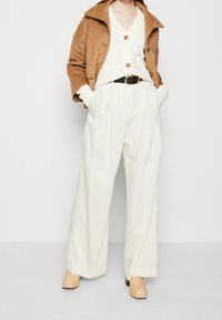 Brun mocka jacka, krämfärgad texturerad tröja, vita randiga vida byxor, svart bälte med runt guldspänne, beige klackskor.