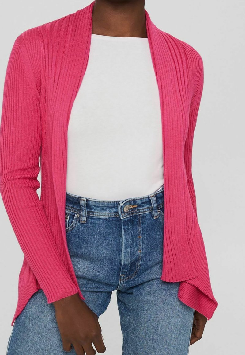 Personne portant un cardigan côtelé rose vif sur un haut blanc avec un jean bleu taille haute, les mains détendues sur les côtés.