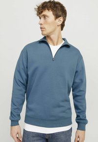 Blue zip-up sweatshirt made of soft material, featuring a stand-up collar, ribbed cuffs, and a relaxed fit, layered over a white t-shirt.