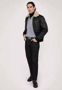 Chaqueta de cuero negro con cuello de borrego, camisa a rayas, pantalones oscuros y zapatos marrones. El modelo está de pie con las manos en los bolsillos frente a un fondo liso.