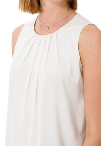 Blouse blanche sans manches avec des plis au niveau de l'encolure. Tissu lisse, léger avec un léger éclat et une coupe décontractée. Collier argenté visible.