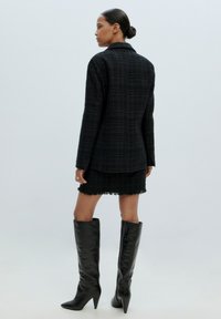 Schwarzer Tweed-Blazer mit Kragen, kombiniert mit einem strukturierten Minirock mit fransigem Saum. Knielange schwarze Leder-Stiletto-Stiefel.