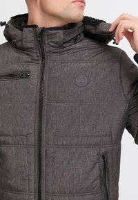 Gray padded jacket with a textured finish. Features a zip closure, side pocket, and hood. Includes a circular logo on the chest.